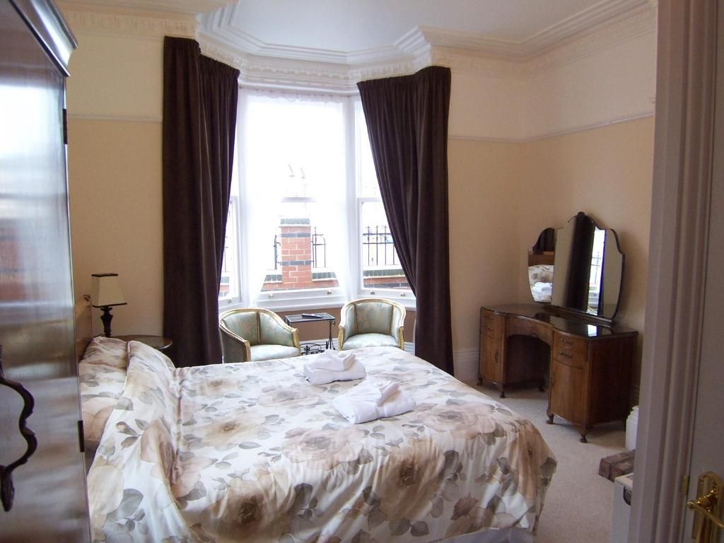 A hotel room with a large bed, two armchairs, a wooden dresser, a mirror, and a window with curtains.