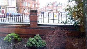 A brick wall with a metal fence, a small garden area with shrubs, and a view of red brick buildings in the background. - The Hollies (Bridlington)
