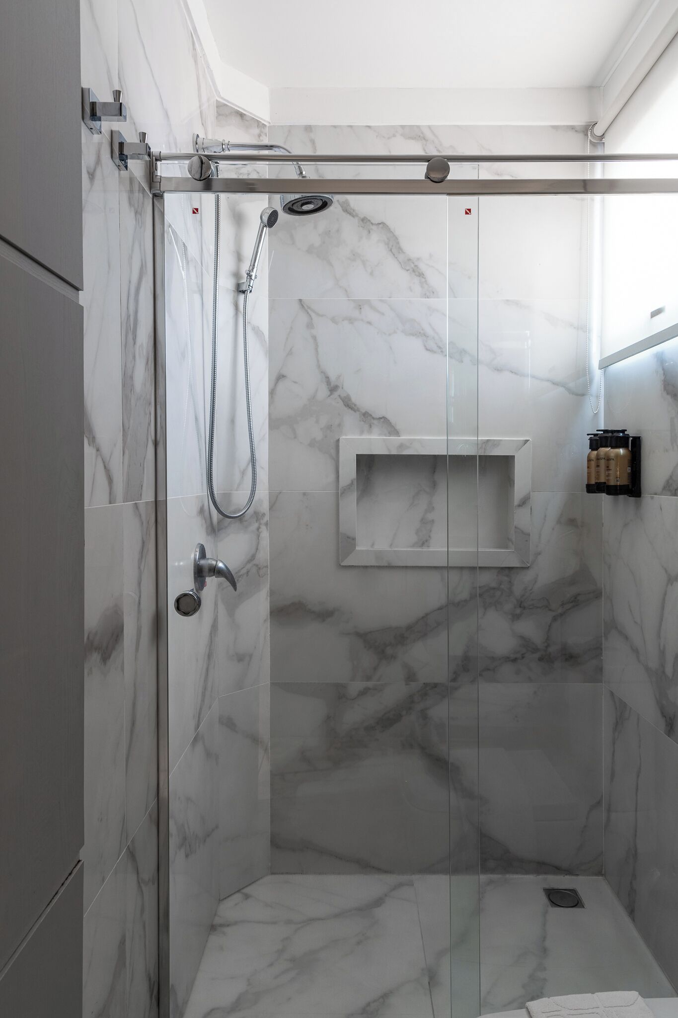 Un box de ducha moderno con puerta de cristal, paredes de mármol y estante integrado.