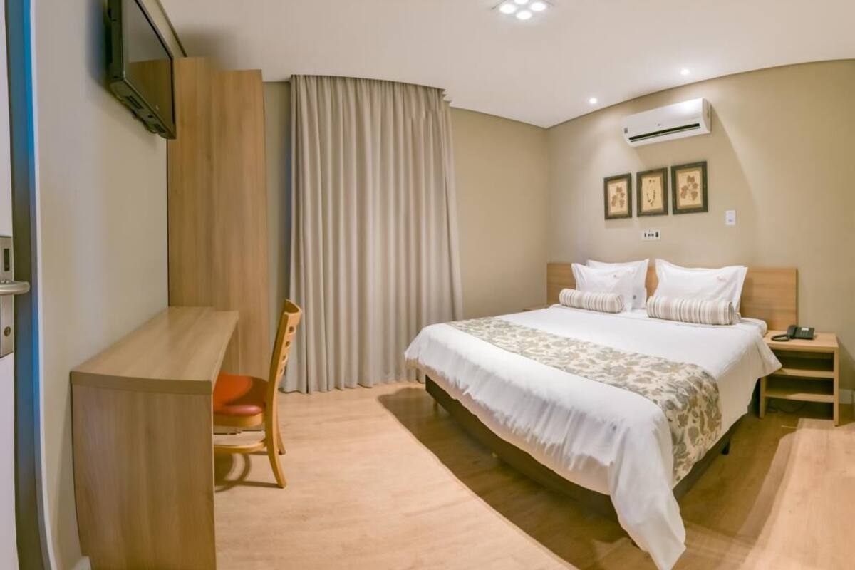 A hotel room with a bed, a desk, a chair, a TV mounted on the wall, and an air conditioning unit.