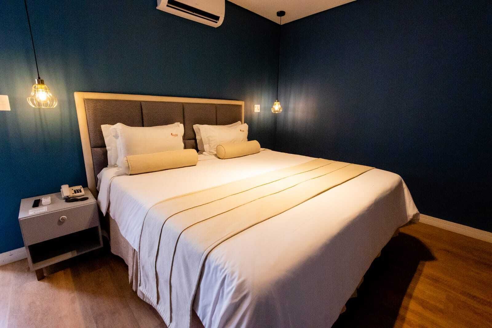 A neatly made bed with white linens and a headboard, a bedside table with a phone and a lamp, and a wall-mounted air conditioner.