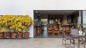 Un espace extérieur aménagé pour les repas, avec plusieurs tables et chaises en bois, une grande plante en pot et une vue sur l’intérieur du bâtiment.
