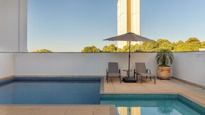 Un espace piscine aménagé sur le toit, offrant une vue dégagée sur un immeuble imposant, avec deux chaises longues et une plante en pot.