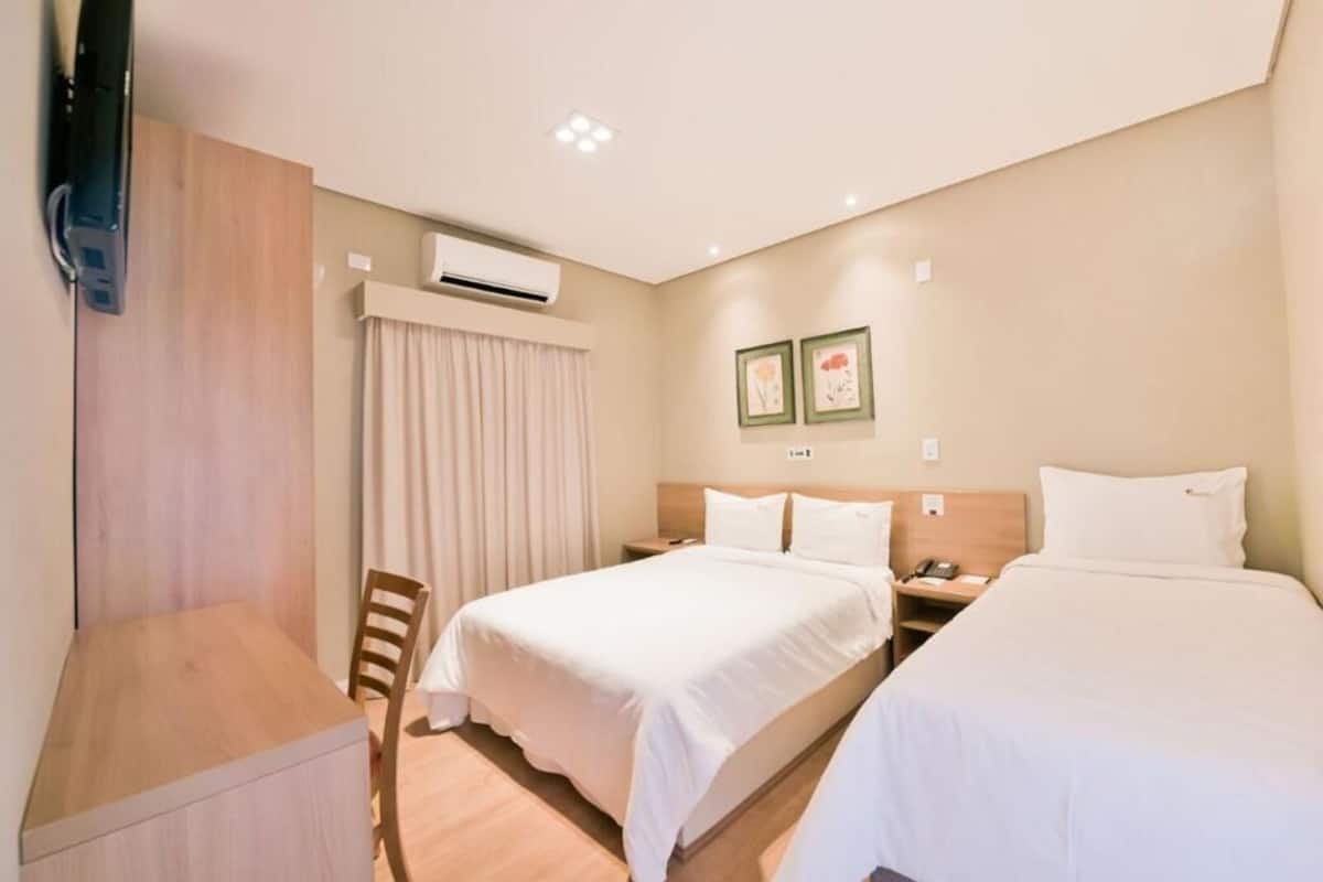 A hotel room with two beds, a desk, a chair, an air conditioning unit, and two framed pictures on the wall.
