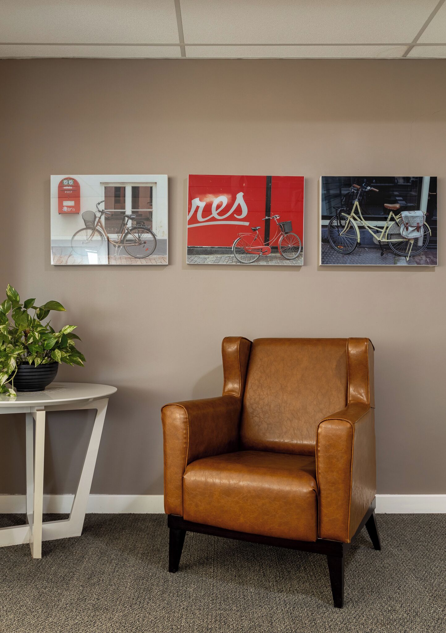 A brown leather armchair in a room with a white table and a potted plant. Three framed pictures of bicycles are on the wall.