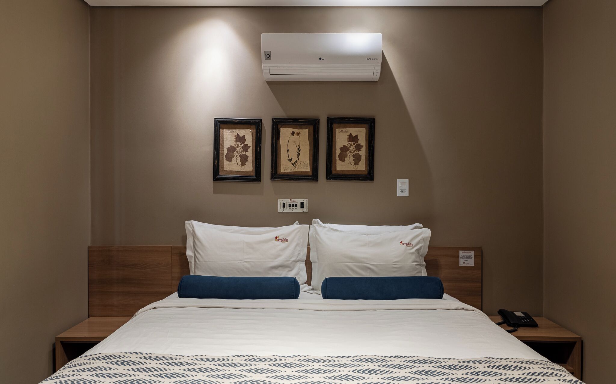 Hotelkamer met een bed, twee kussens, een nachtkastje met een telefoon en drie ingekaderde botanische prints aan de muur.