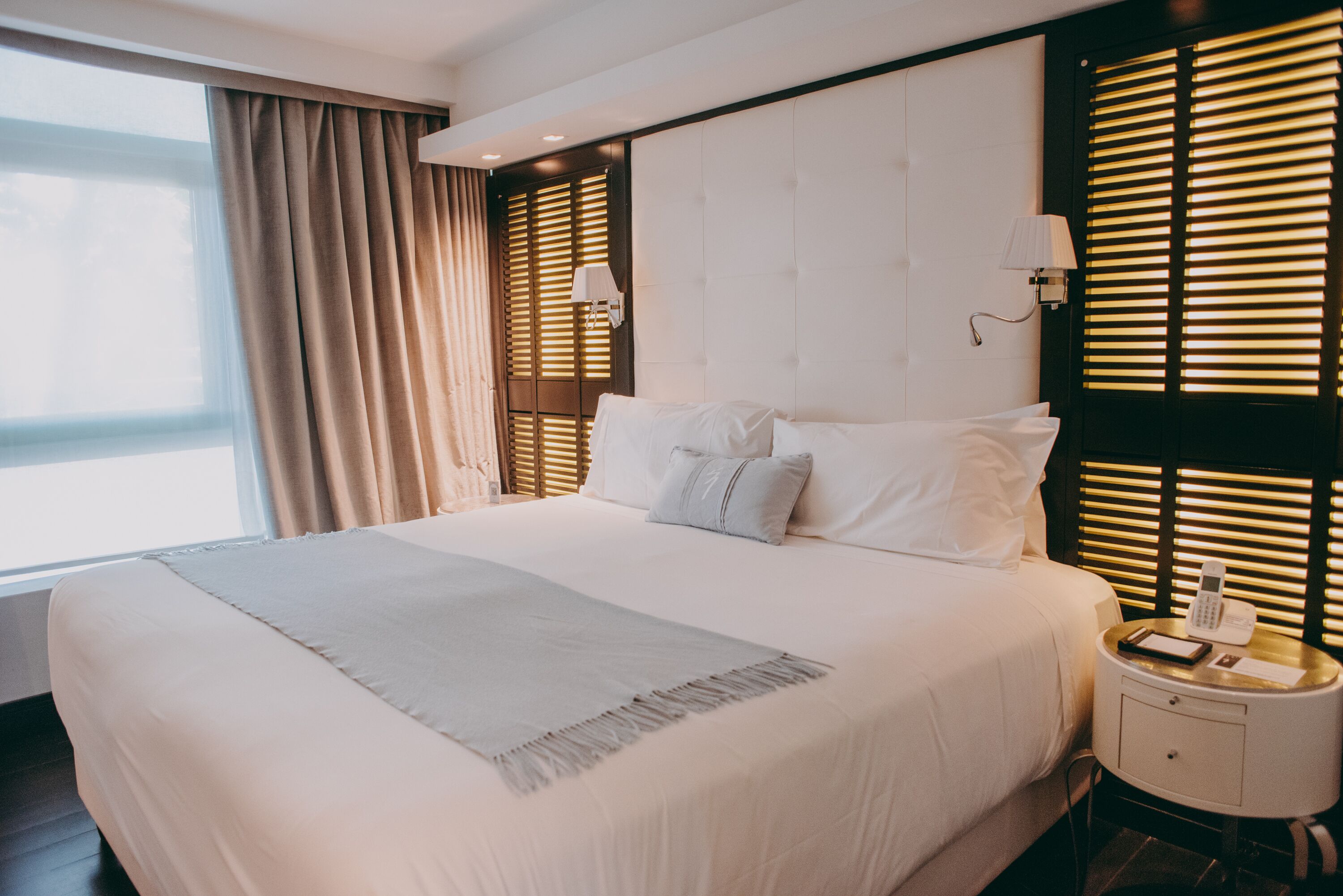 A hotel room with a large bed, a bedside table with a lamp, and a window with blinds.