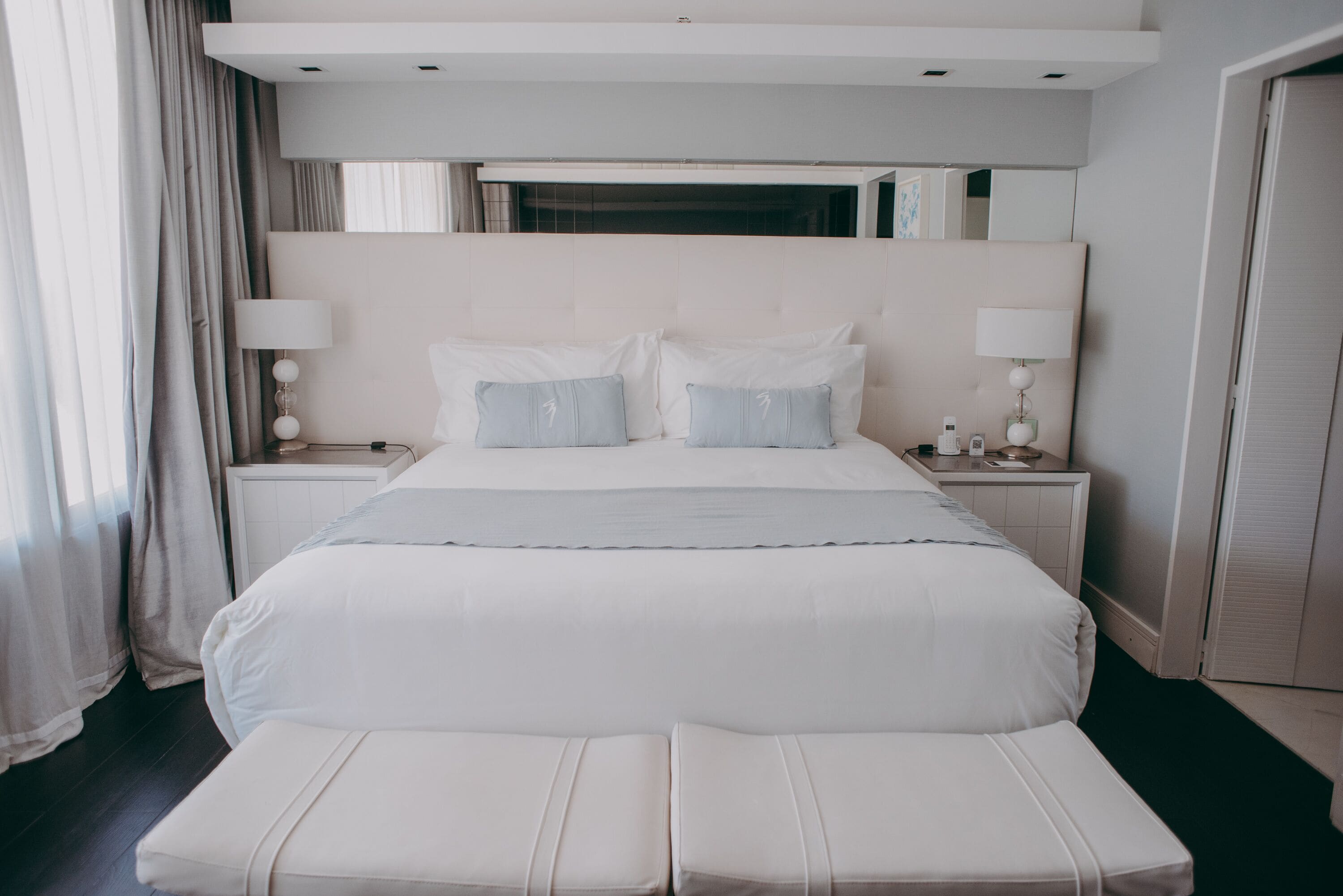 A modern hotel bedroom with a large bed, two bedside tables with lamps, a headboard, and a view of the outside through a window.