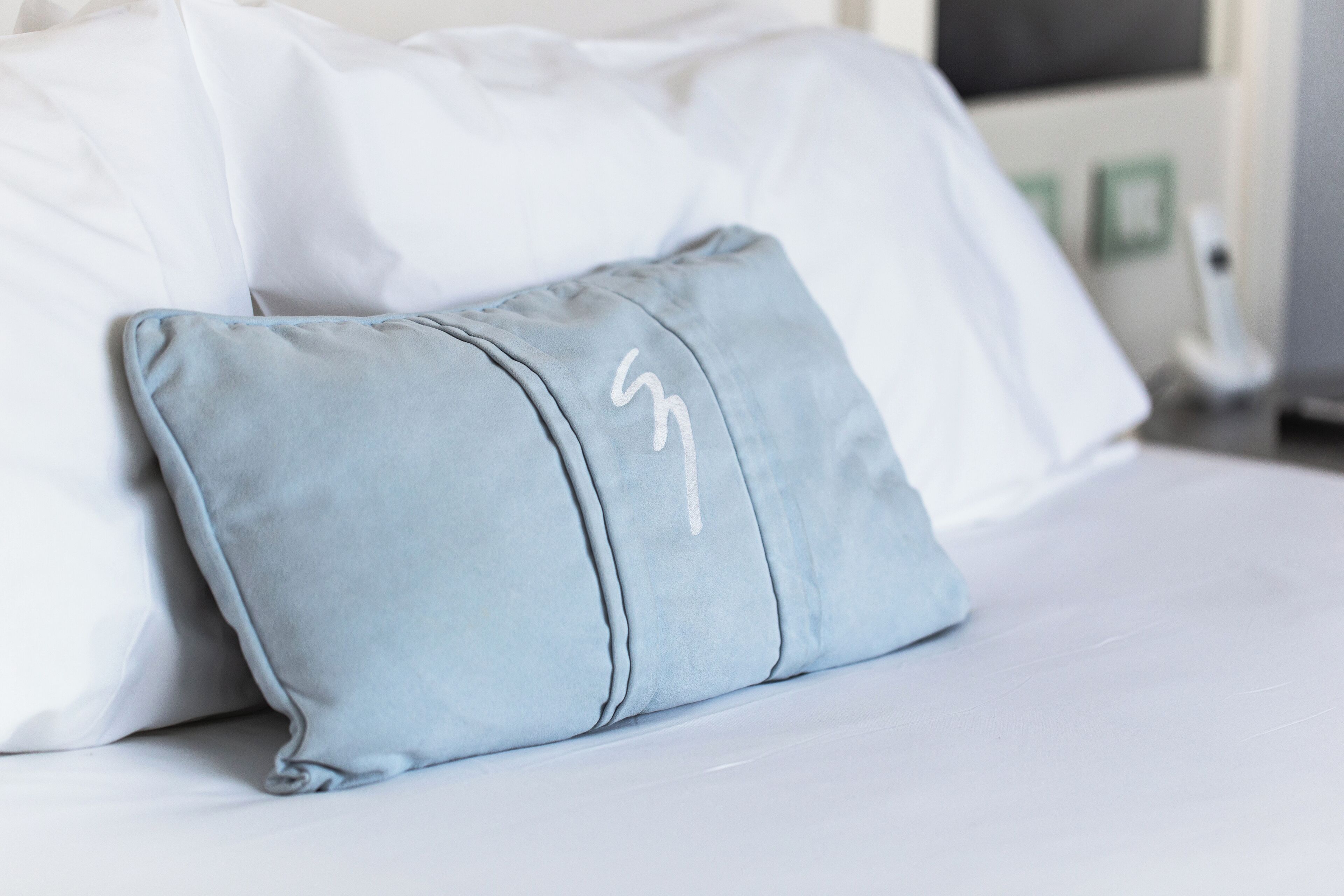 A bed with white bedding and a light blue pillow with a white design.