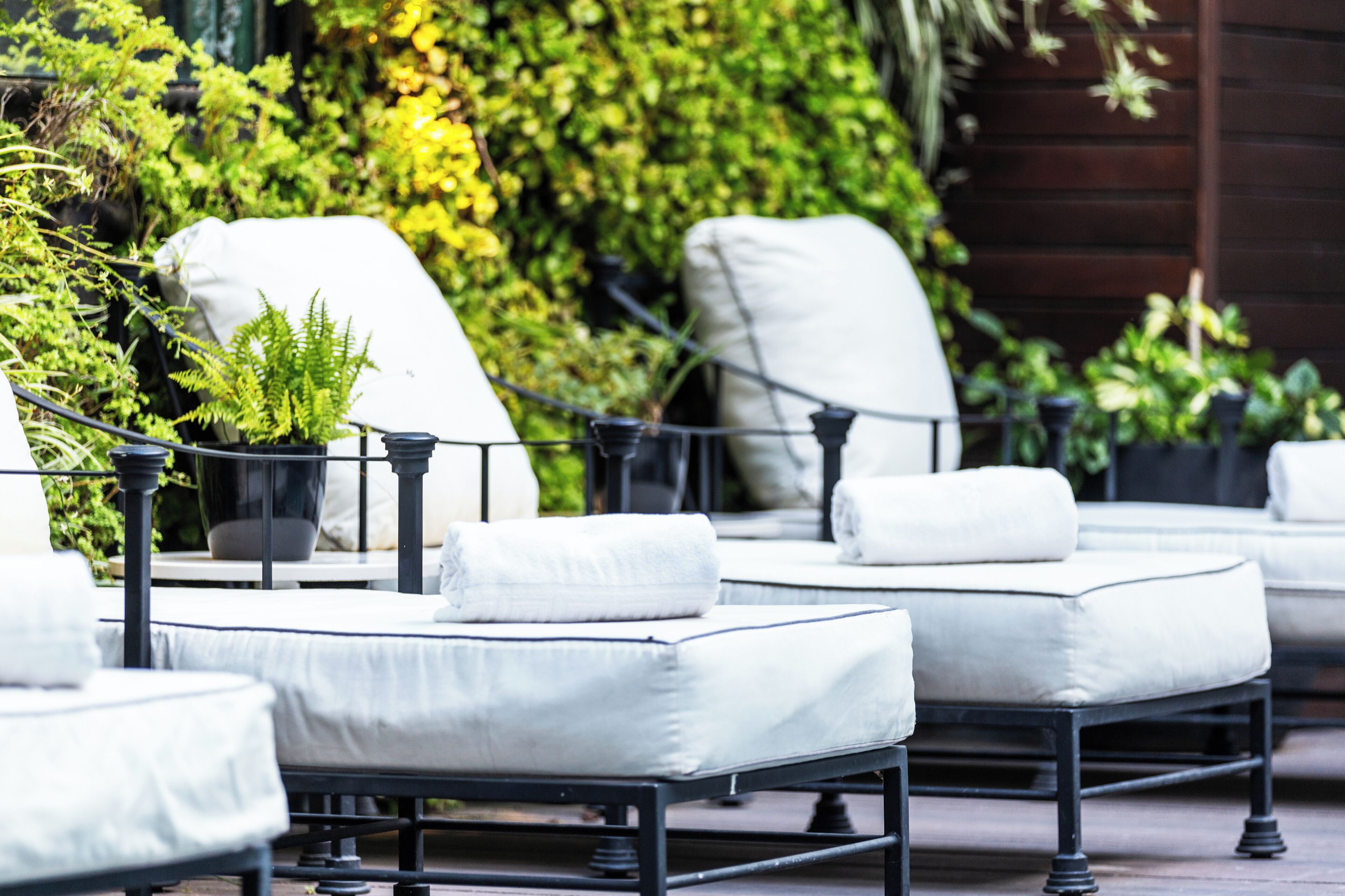 A patio area with white cushioned chairs and tables, surrounded by lush greenery and a wooden fence.