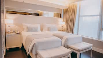 A hotel room with two beds, bedside tables with lamps, a large window with curtains, and a wooden floor.
