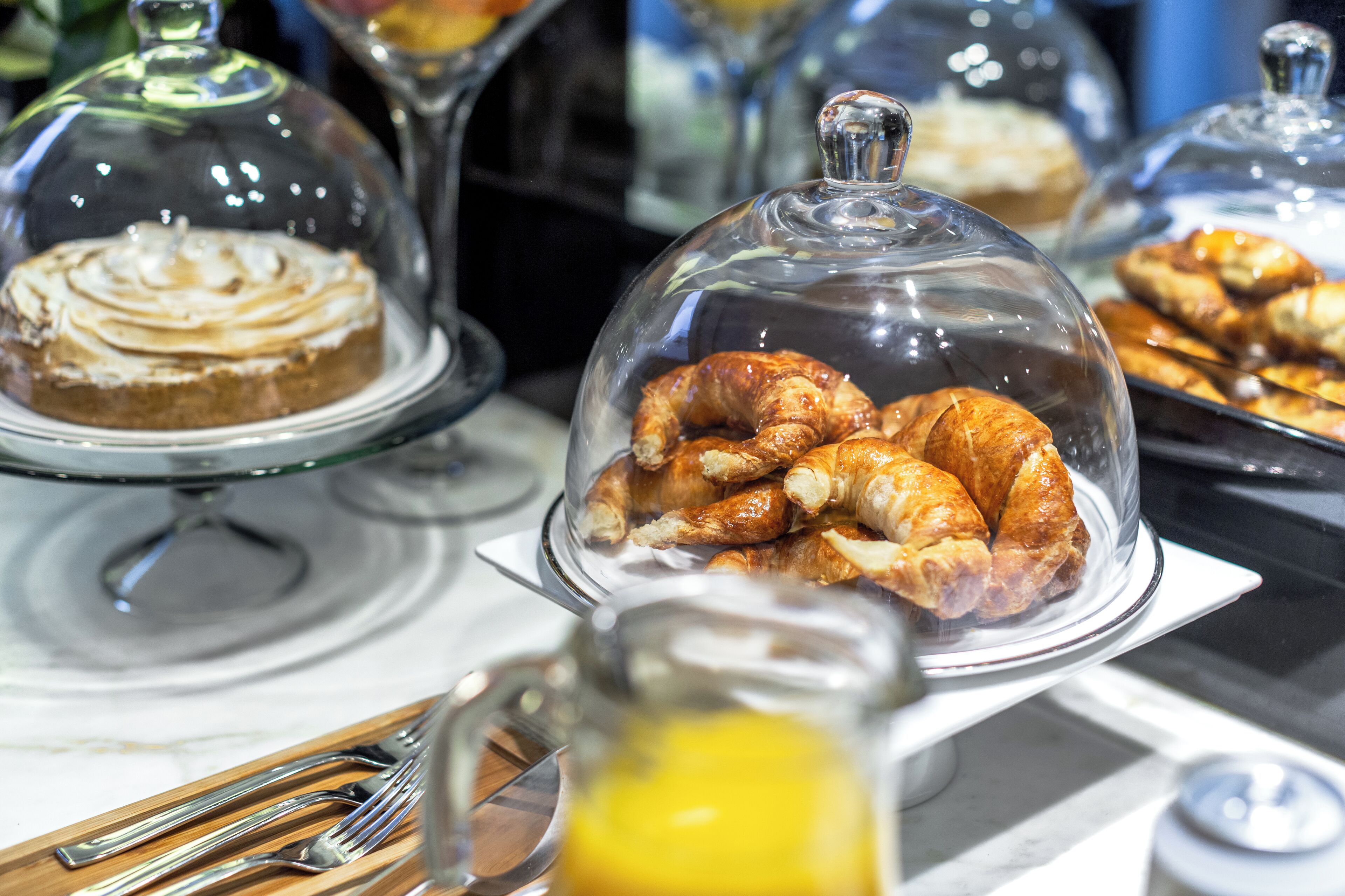 Ein Buffet mit verschiedenen Backwaren unter Glaskuppeln, darunter Croissants und ein Kuchen.