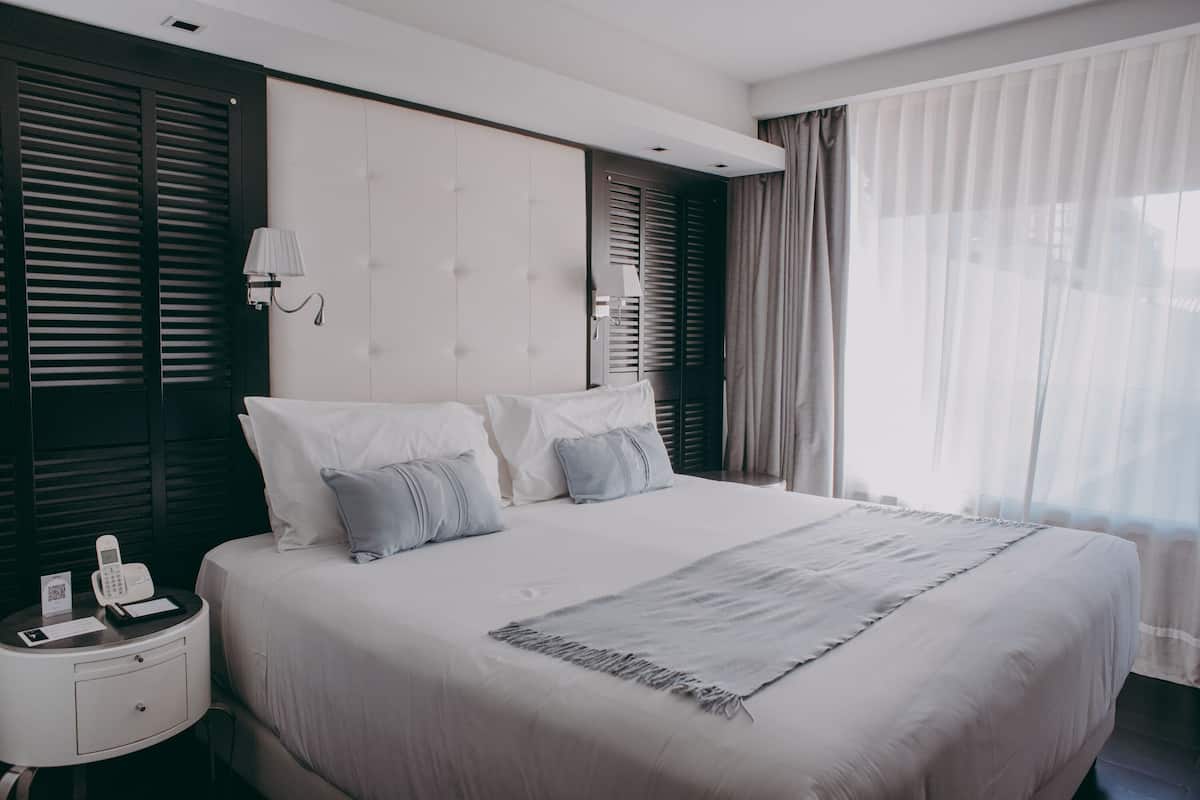 A neatly made bed with white linens and a grey throw, a bedside table with a lamp, and a phone.