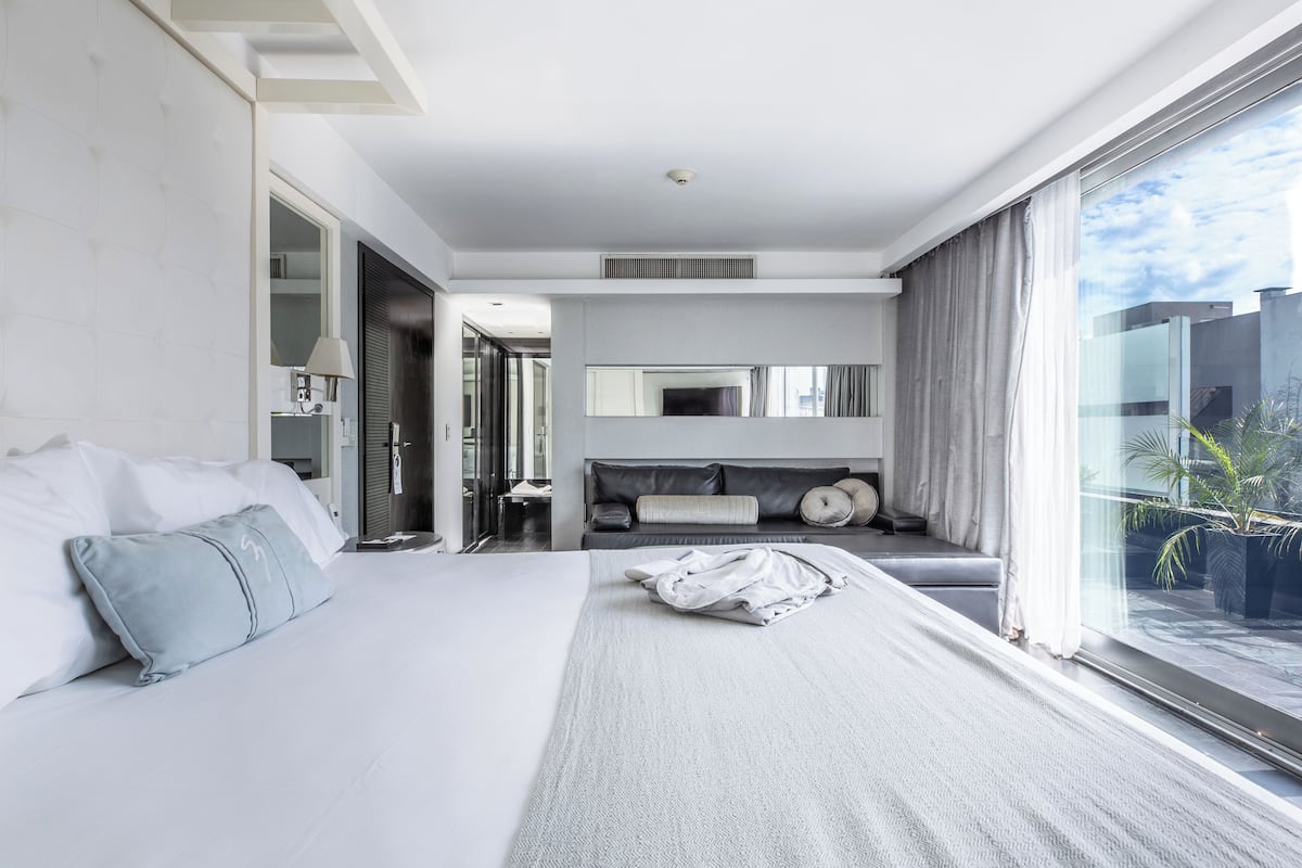 A modern hotel room with a large bed, a sofa, a TV, and a large window with a view of the city.