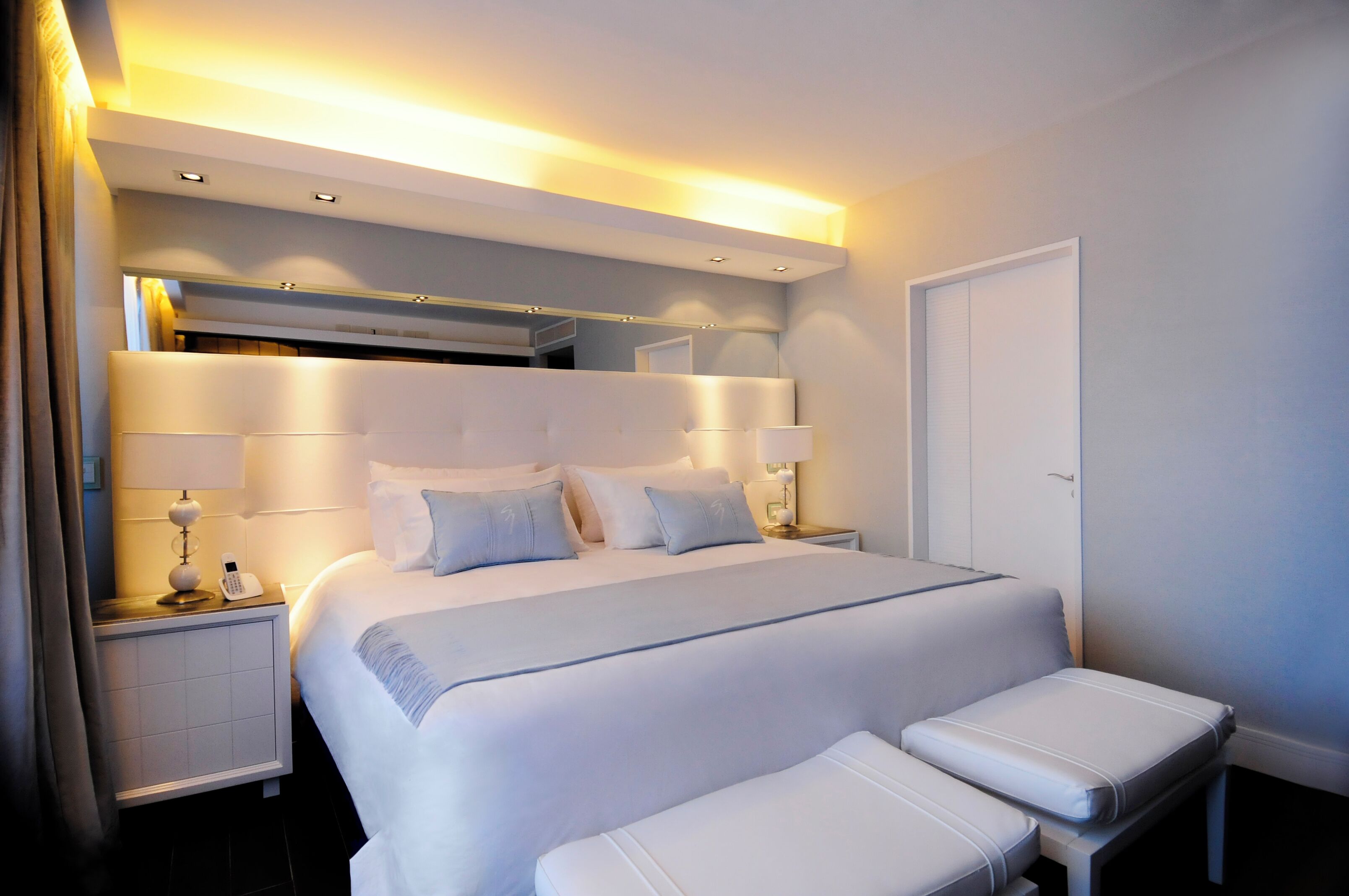 A modern hotel room with a large bed, bedside tables, a bench, and a door.