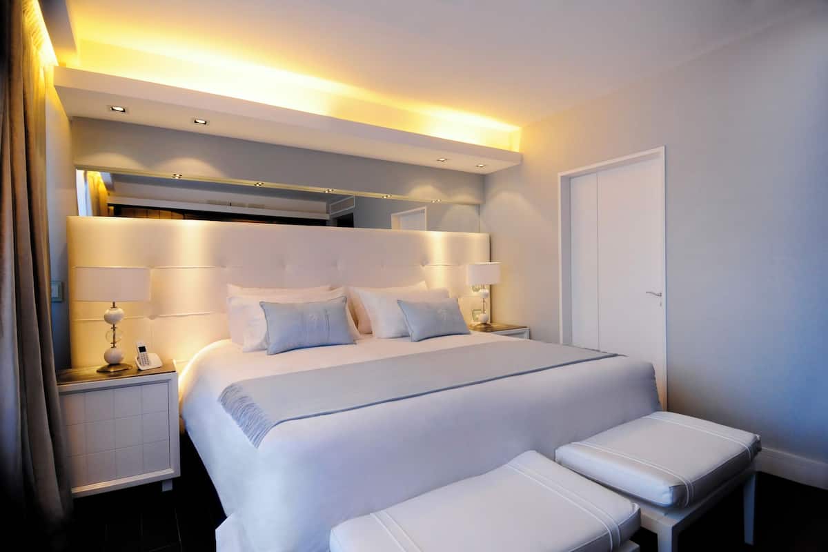 A modern hotel room with a large bed, bedside tables, a bench, and a door.