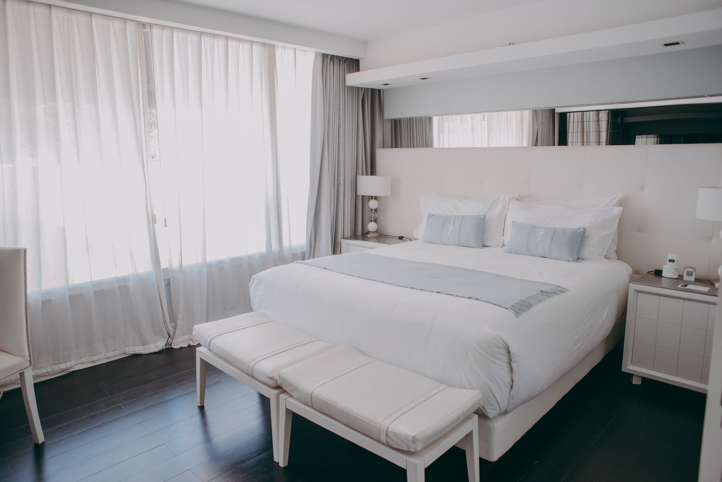 Una habitación de hotel moderna con una cama grande, un banco, una mesita de noche y un amplio ventanal con cortinas.
