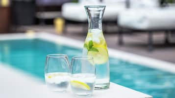Escena junto a la piscina con una jarra de vidrio transparente y dos vasos llenos de un líquido claro, adornados con rodajas de limón, sobre una superficie blanca.