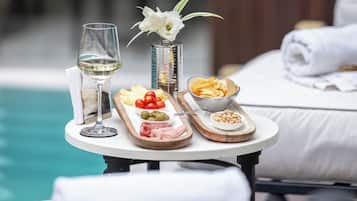 A table with a glass of white wine, a small vase with white flowers, and a wooden board with various snacks and a bowl of olives.
