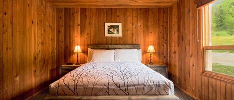 A wooden-paneled bedroom with a bed, two bedside lamps, and a window with a view of greenery.