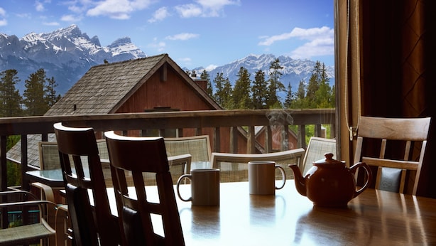 A wooden table with chairs and a teapot on a balcony overlooking snow-capped mountains and a wooden cabin. - Banff Gate Mountain Resort (Canmore)