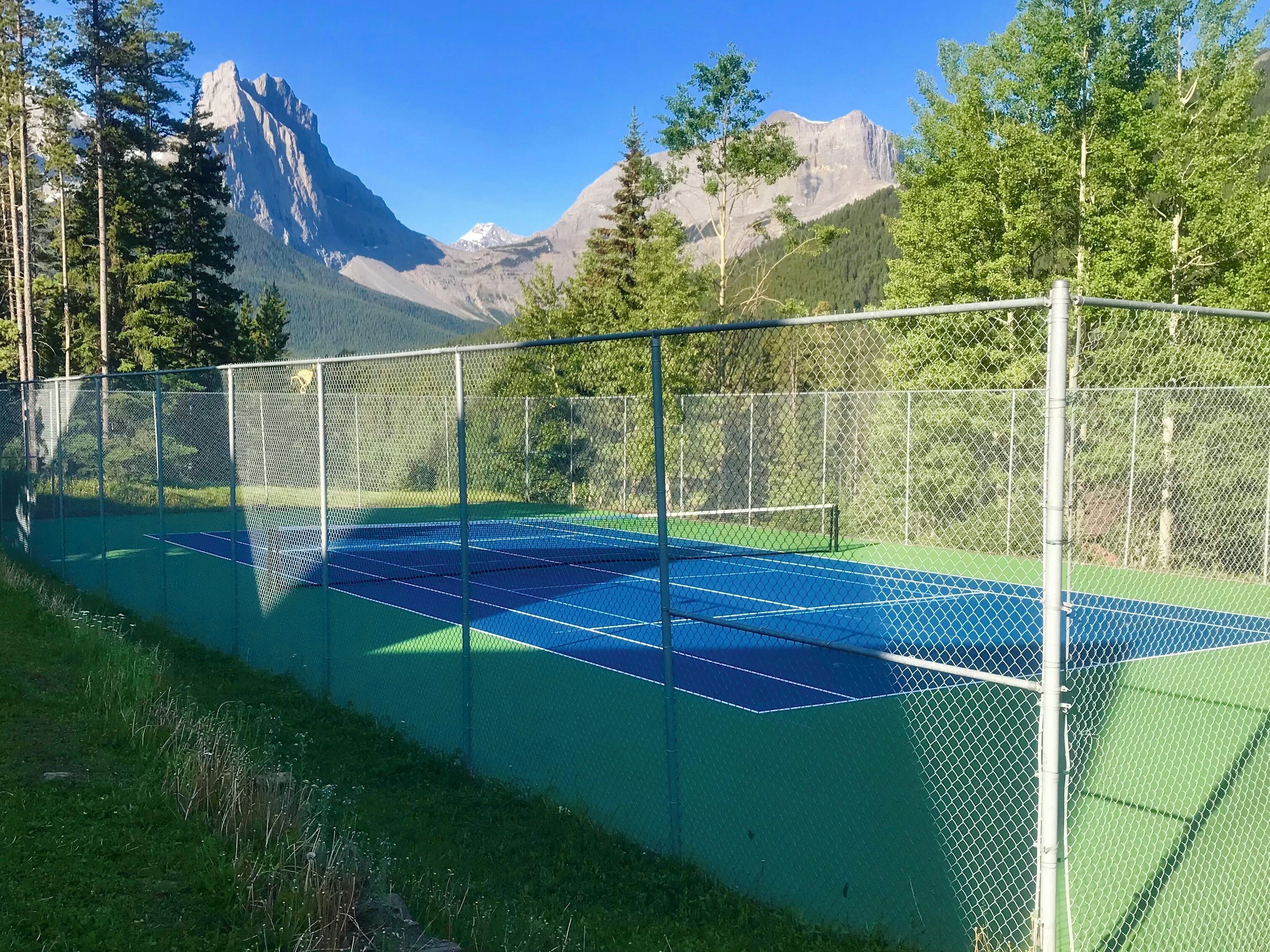 A tennis court with a chain-link fence, surrounded by trees and mountains in the background.