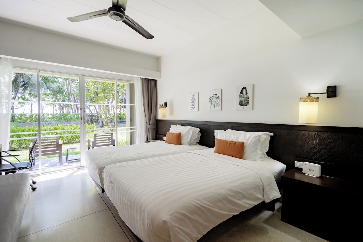 Une chambre d’hôtel avec deux lits, un ventilateur de plafond, un bureau avec une chaise et une vue sur la verdure grâce à de grandes fenêtres.