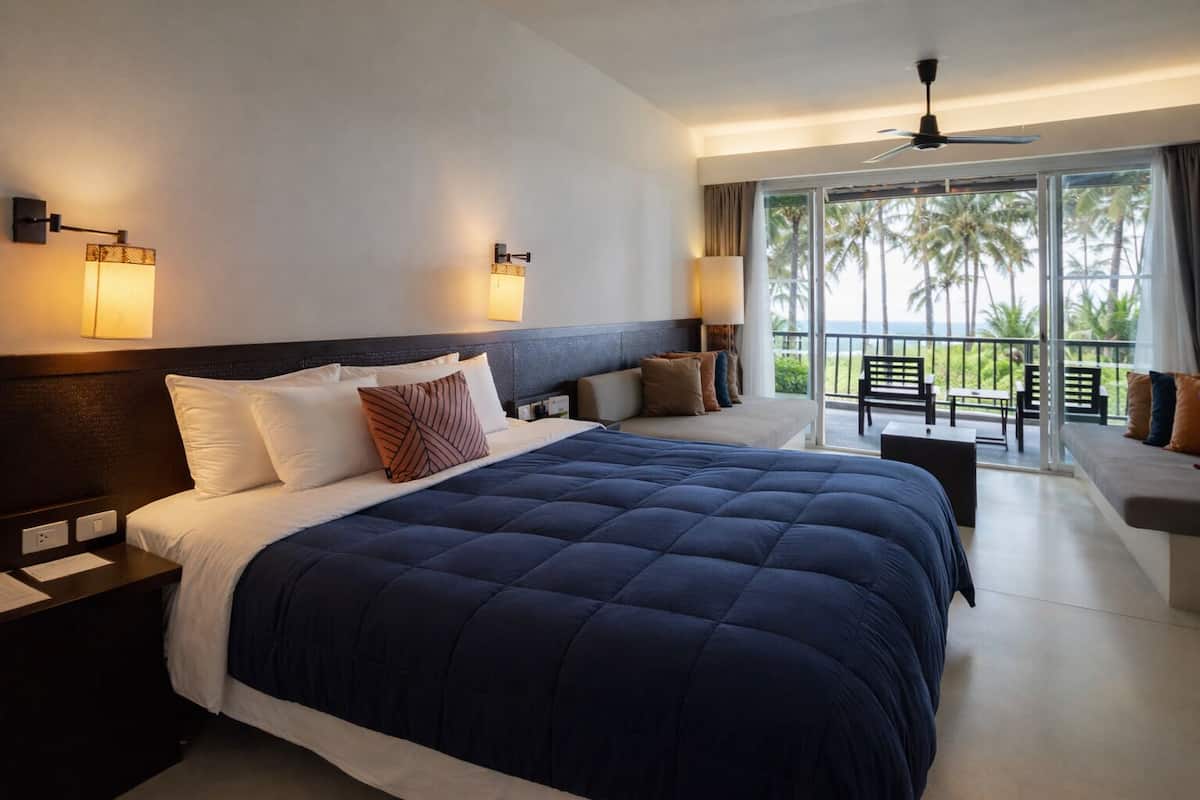 Ein Hotelzimmer mit einem großen Bett, einem Balkon mit Sitzgelegenheit und Blick auf Palmen.