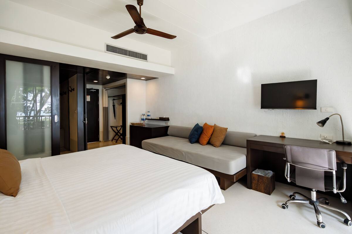 Ein Hotelzimmer mit Bett, Sofa, Schreibtisch, Stuhl, Fernseher und Deckenventilator.