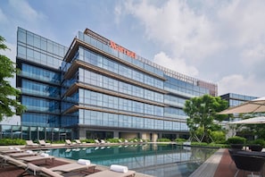 Un hôtel Marriott doté d’une façade moderne en verre, d’un espace piscine sur le toit avec des chaises longues, et surplombant un ciel d’un bleu éclatant.