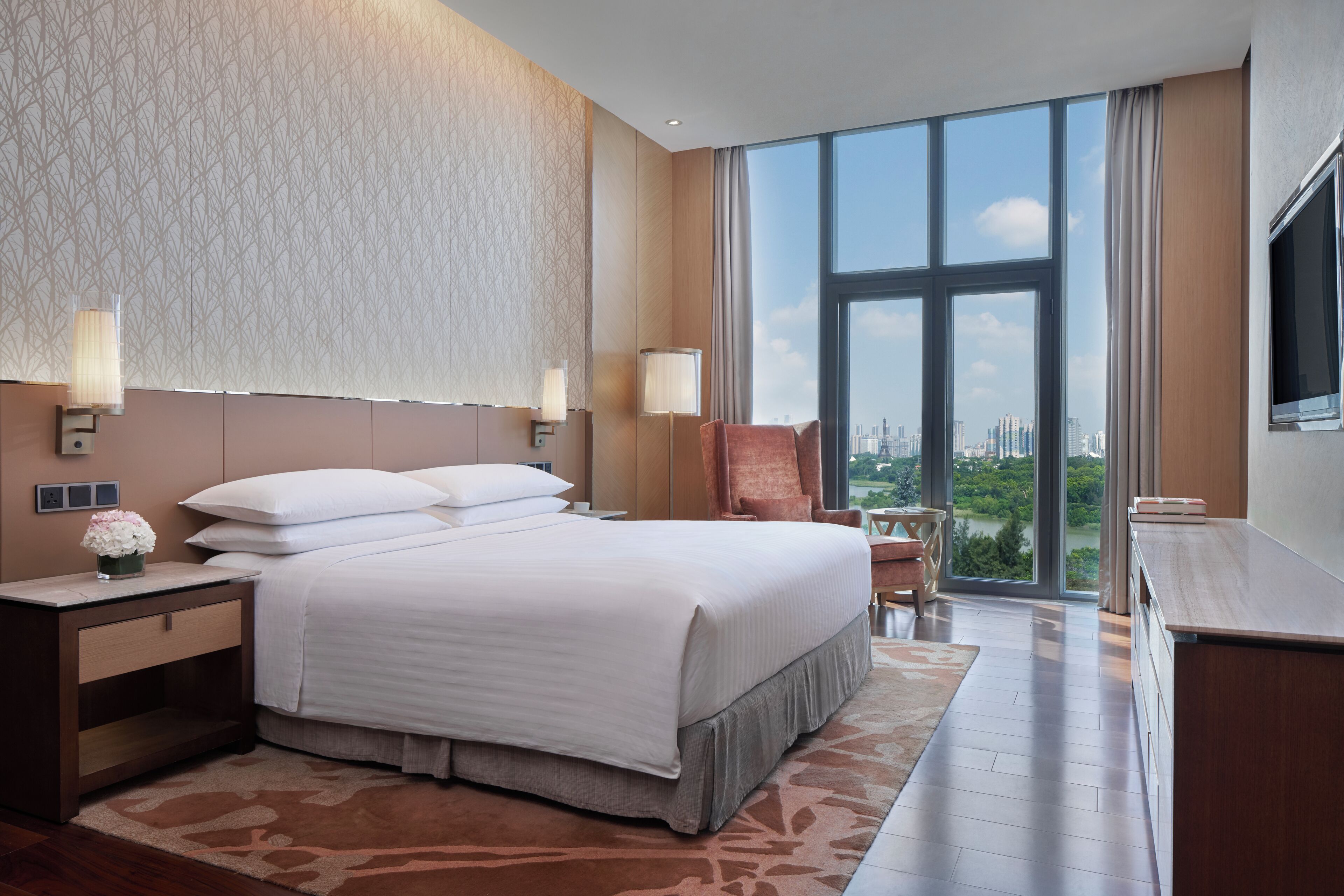 A hotel room with a large bed, a nightstand, a chair, a desk, and a large window offering a city view.