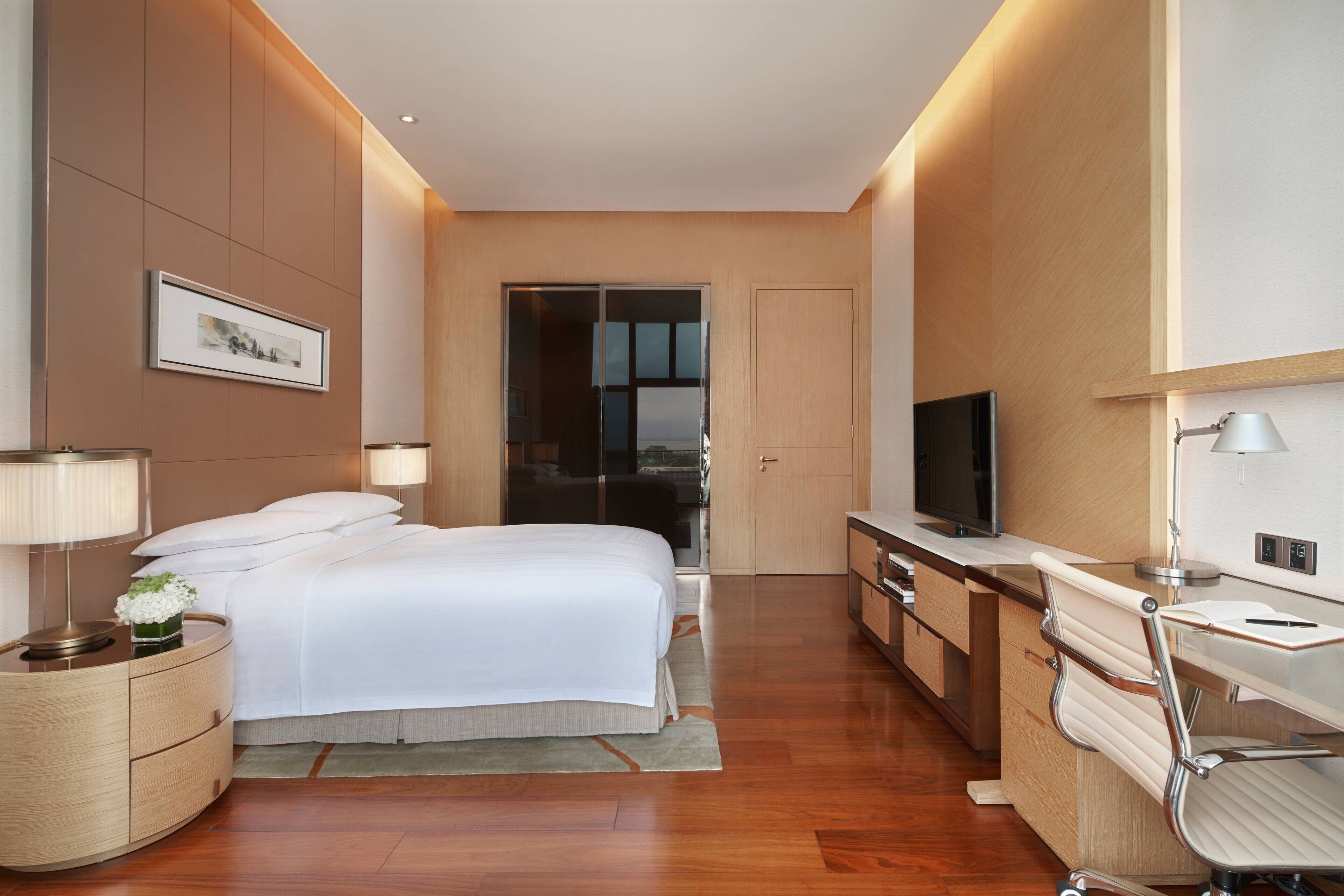 A modern hotel room with a large bed, bedside tables, a desk with a chair, a television, and a mirror.
