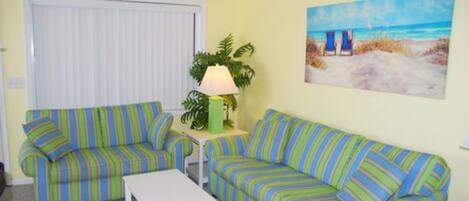 A living room with two striped sofas, a small table, a lamp, and a painting of a beach scene.