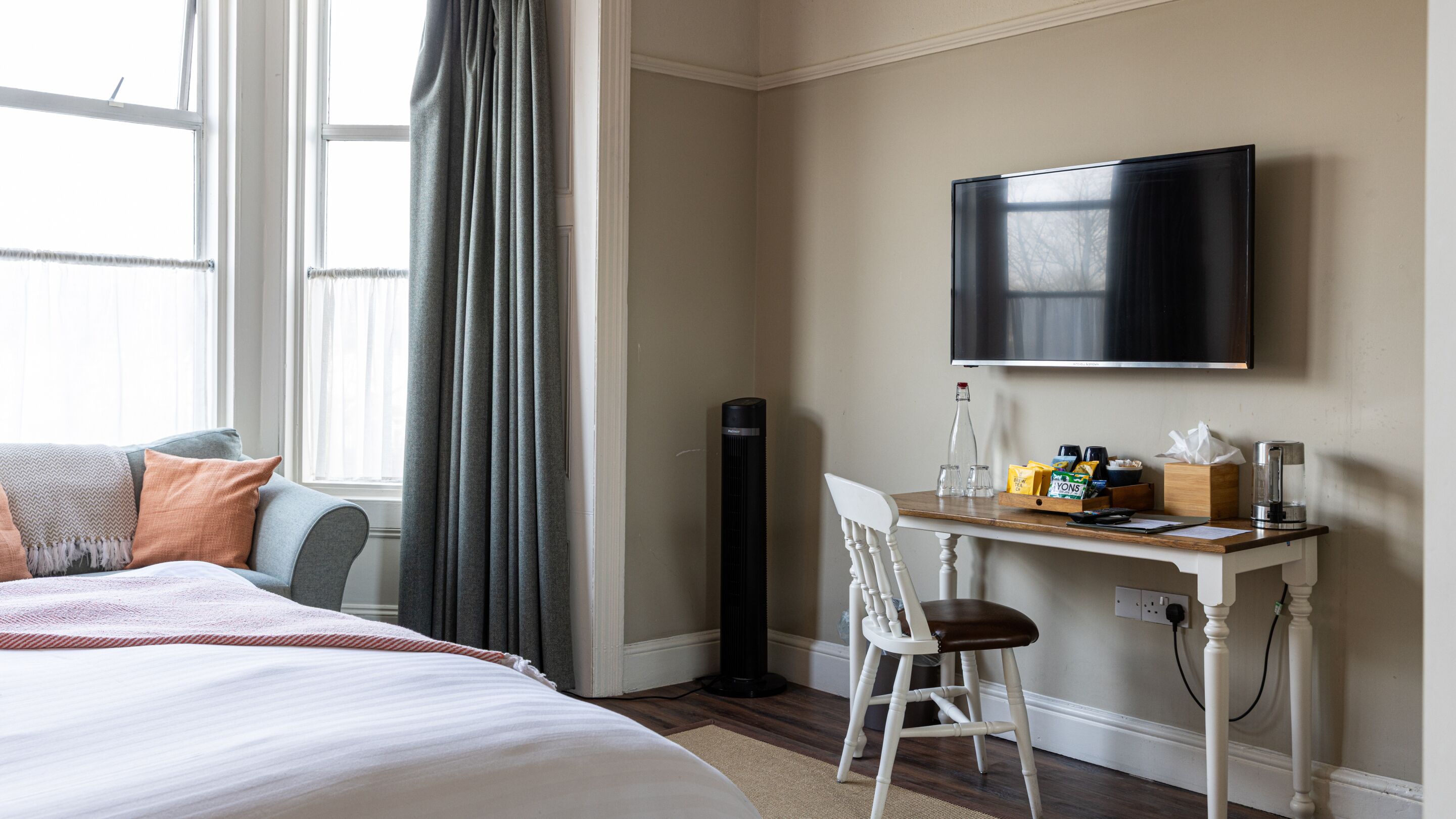 A hotel room with a bed, a desk with a chair, a flat-screen TV, and a window with curtains.