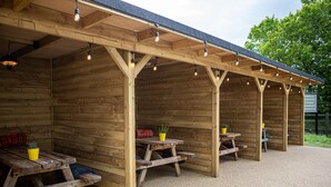 A covered outdoor seating area with wooden benches and picnic tables, featuring overhead lighting and potted plants. - Burrator Inn (Yelverton)
