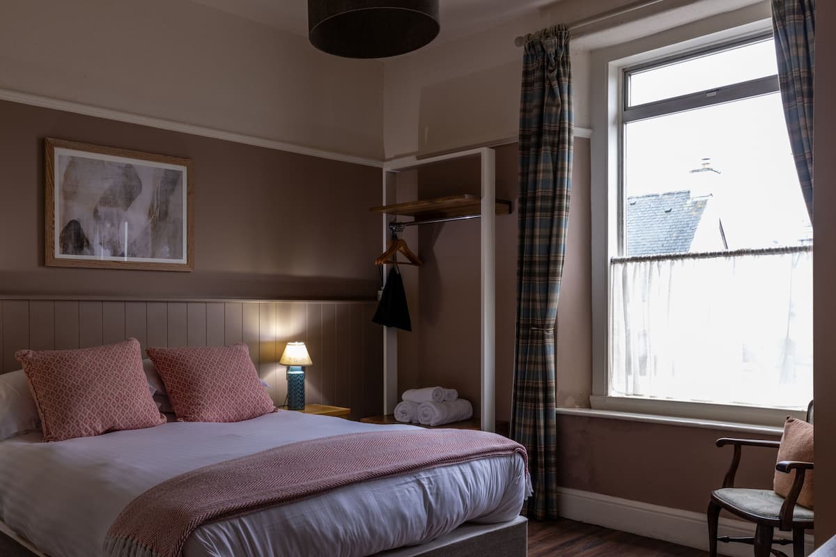A bedroom with a bed, two bedside lamps, a framed picture on the wall, a window with curtains, and a chair.
