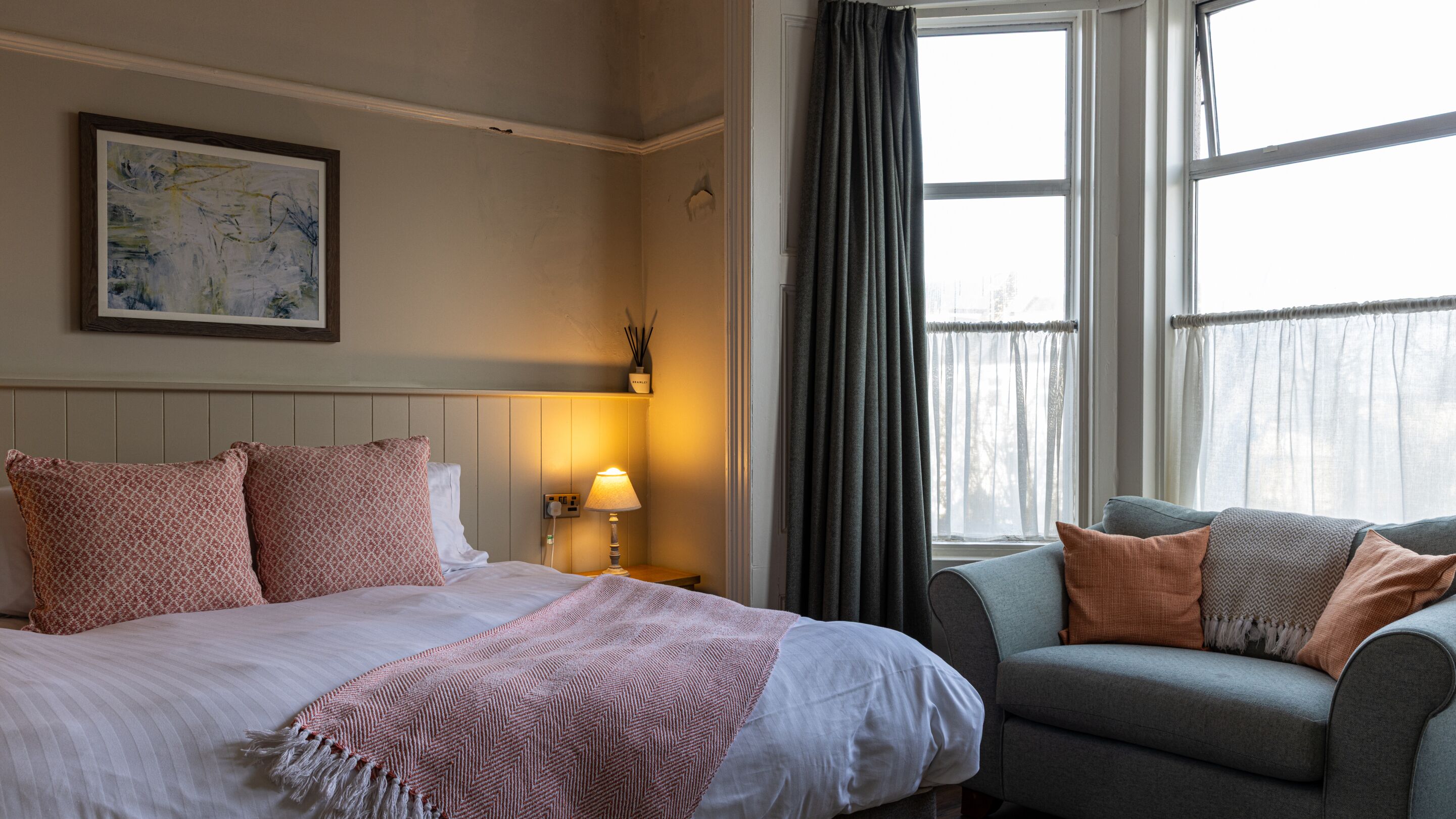 A hotel room with a bed, a bedside table with a lamp, a chair, and a window with curtains.