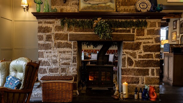 A stone fireplace with a mantel decorated with greenery, a lit candle, and a rustic wooden chair with a cushion. - Burrator Inn (Yelverton)