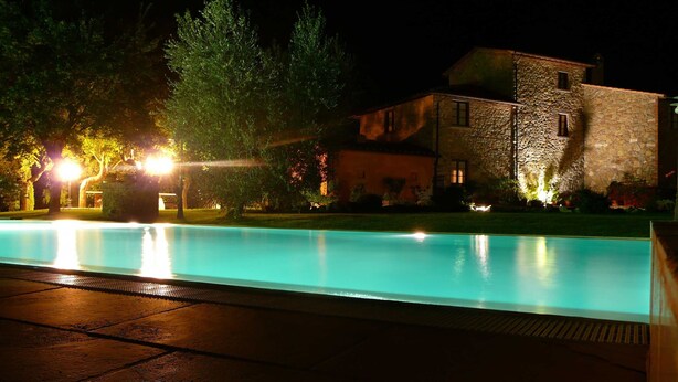 Una piscina nocturna con edificios de piedra al fondo.