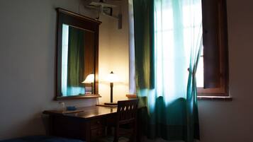 A room with a bed, a desk with a lamp, a chair, a mirror, and a window with curtains.