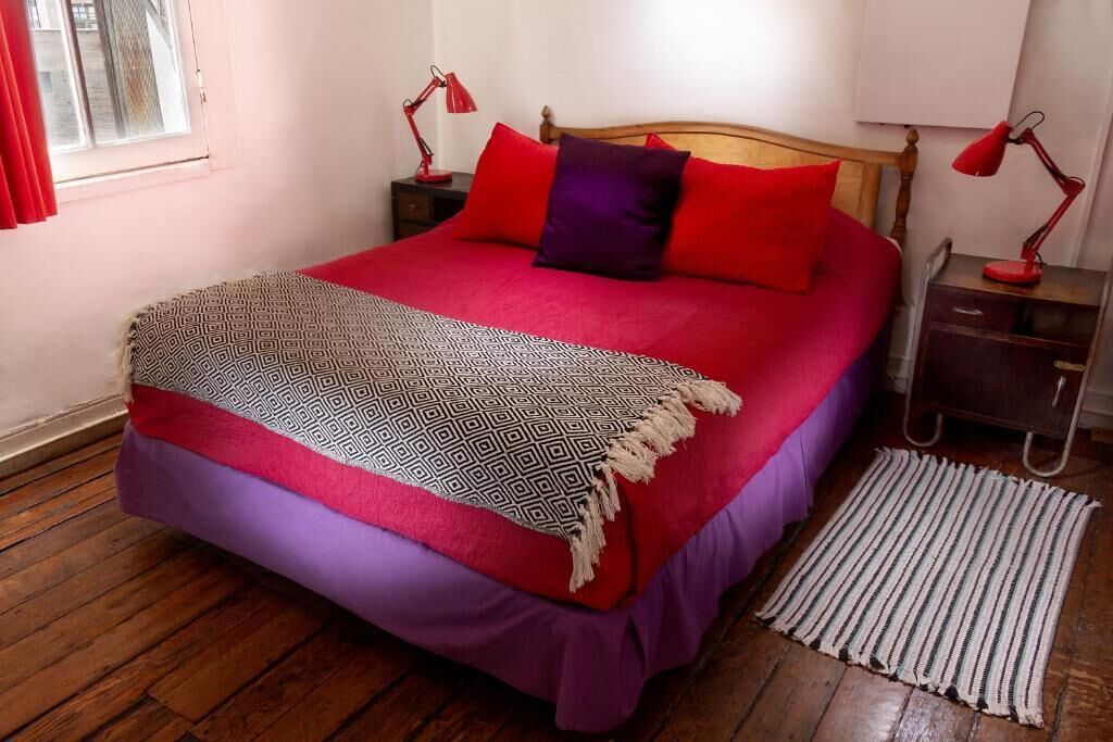 Ein Bett mit einer gemusterten Decke, roten und violetten Kissen sowie einem Holz-Kopfende. Es gibt zwei Nachttische mit Lampen.