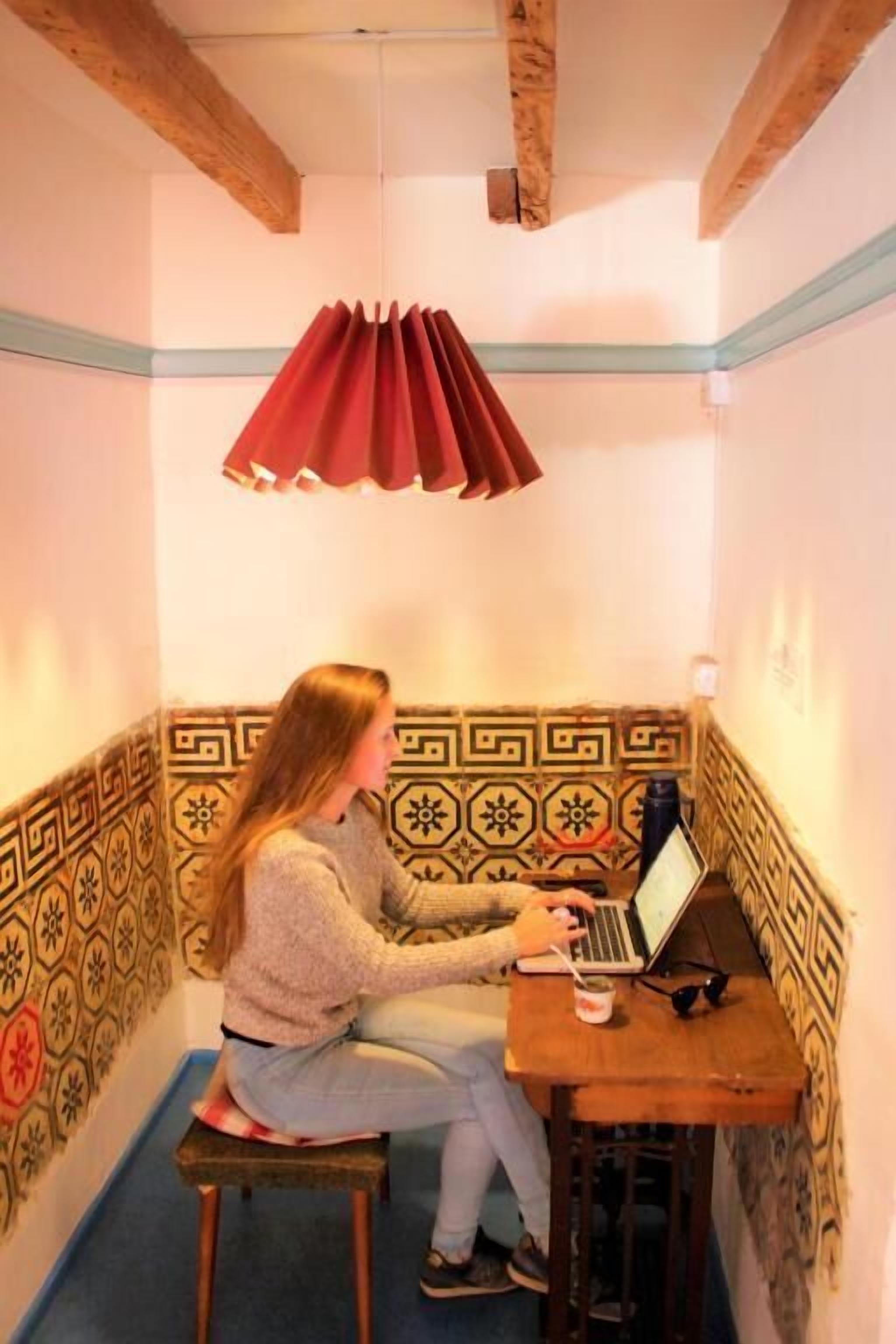 A person sitting on a stool at a wooden table with a laptop, in a room with patterned walls and exposed wooden beams.