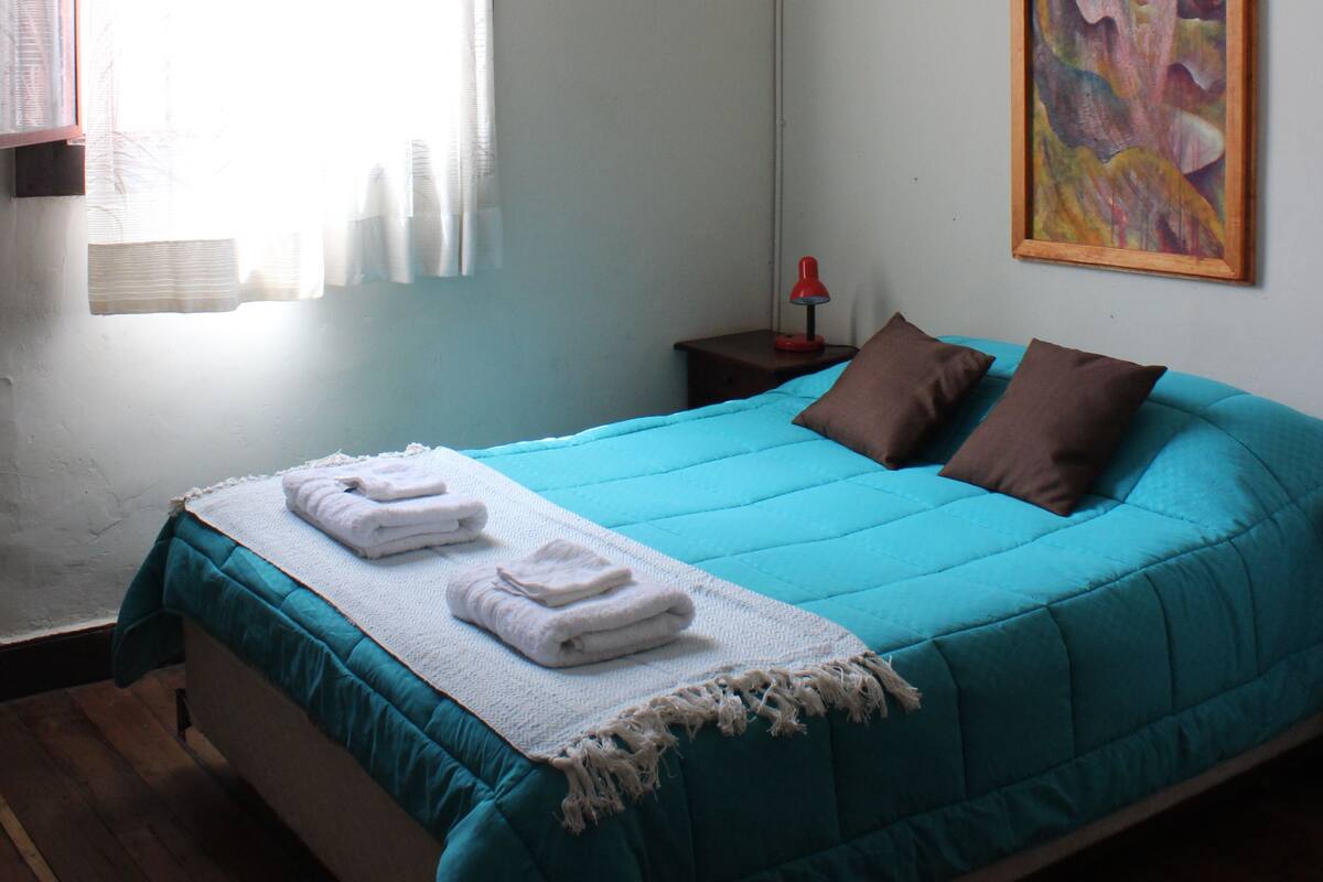 Une chambre à coucher avec un lit, une table de chevet, une lampe, un tableau et une fenêtre avec des rideaux.