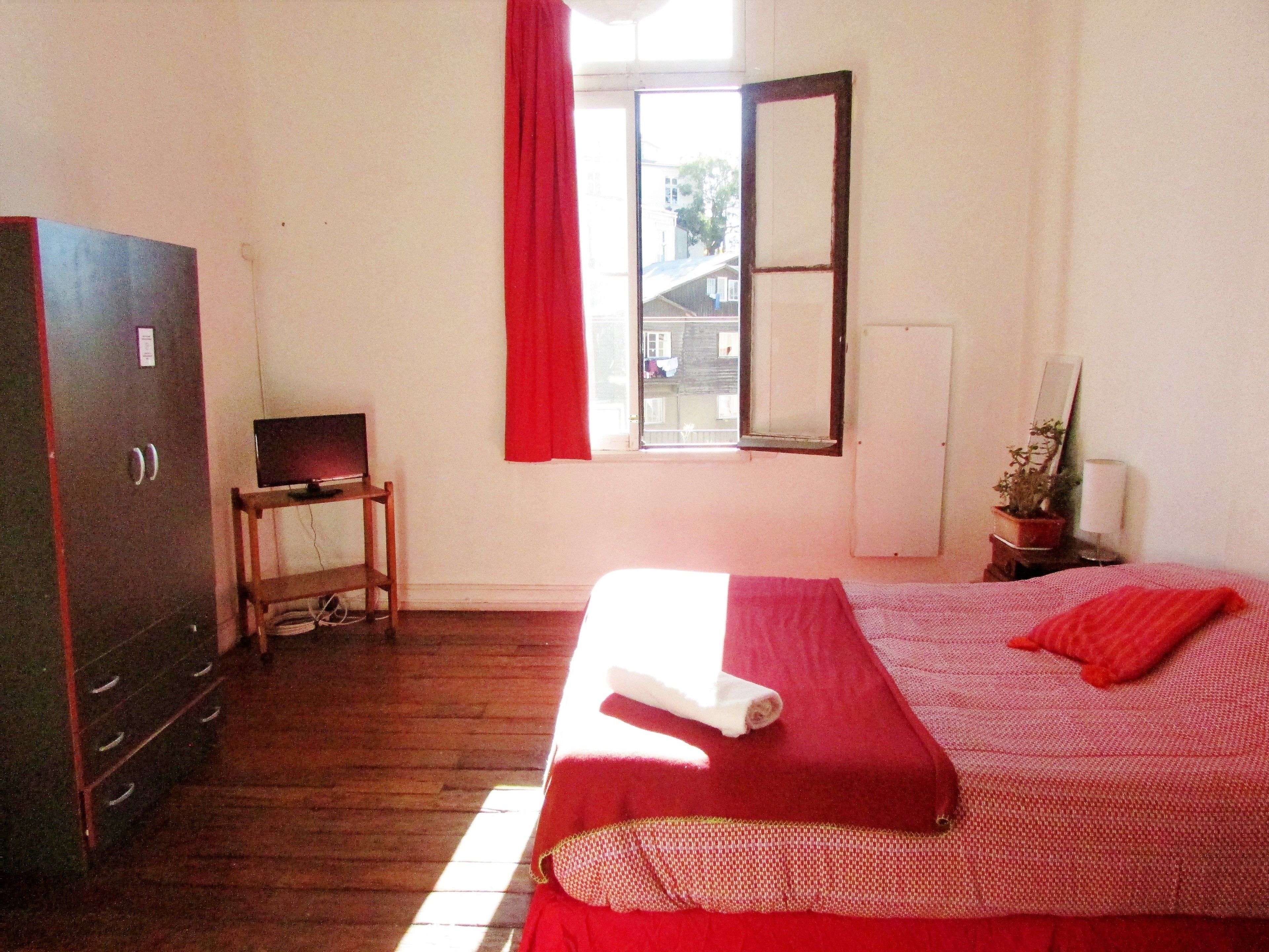 A room with a bed, a desk, a wardrobe, a window with red curtains, and a potted plant.