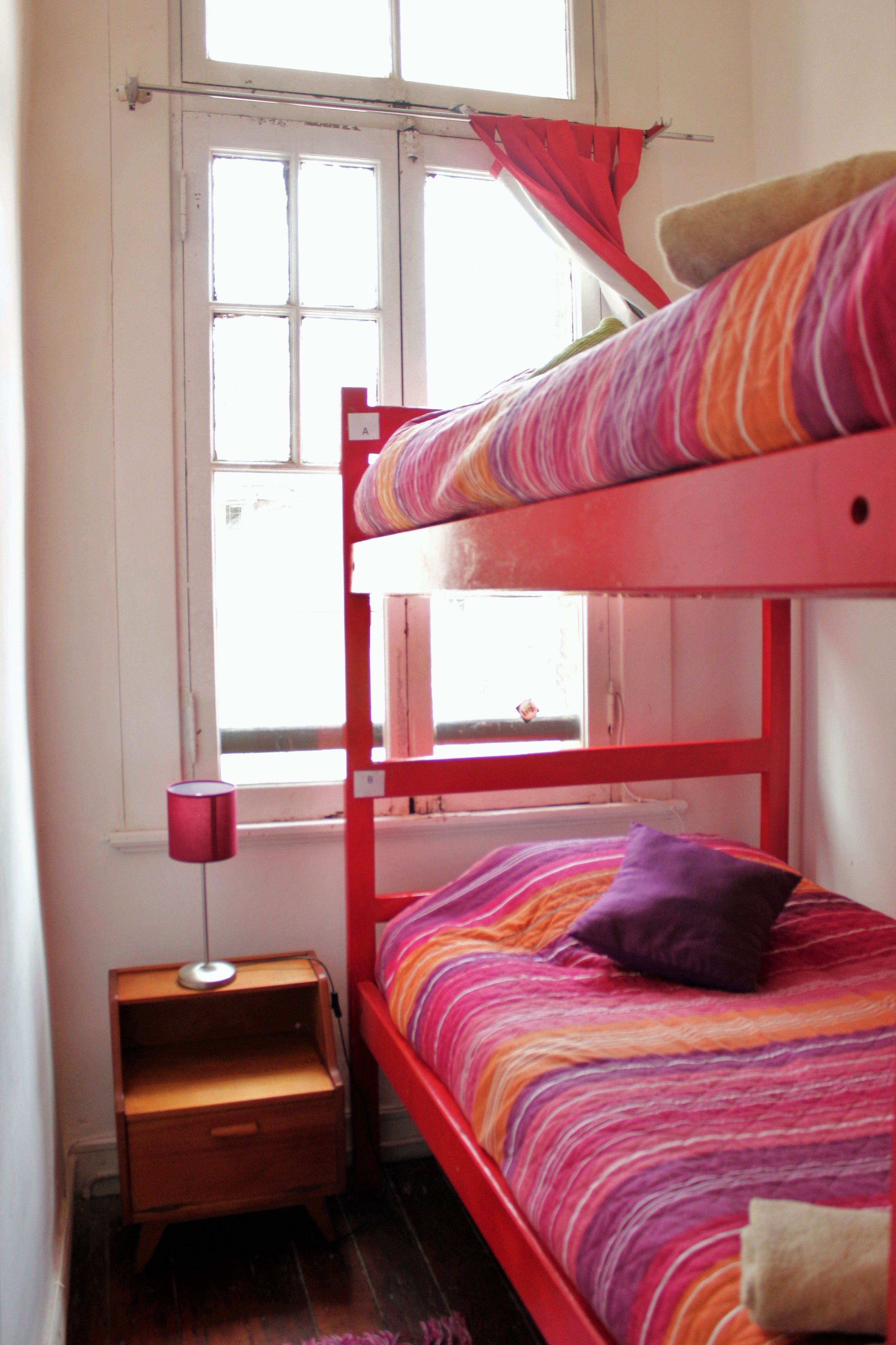 A bunk bed with a colorful striped bedspread and a purple pillow. There is a small wooden nightstand with a lamp beside the bed. A window with a red curtain is visible in the background.