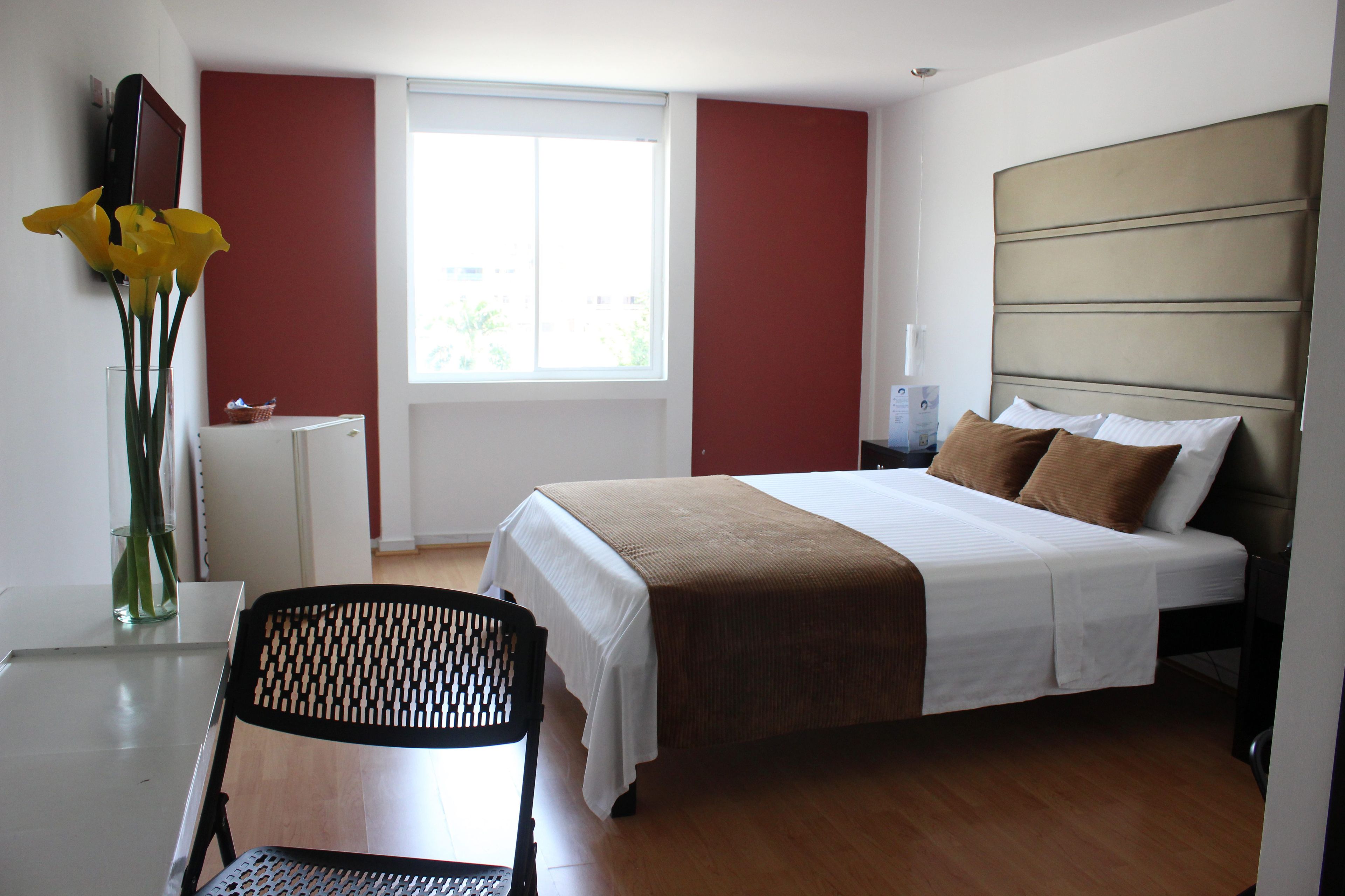 A hotel room with a bed, a desk, a chair, a vase with flowers, and a window.