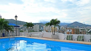 A swimming pool with lounge chairs and a lamp, surrounded by palm trees and a cityscape with mountains in the background. - Hotel Metropolis Plaza (Cali)