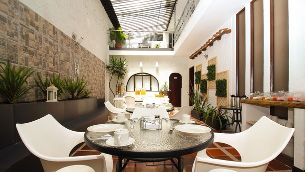 Un comedor con una mesa redonda dispuesta para cuatro personas, rodeada de sillas blancas, en un patio con pared de ladrillo y plantas en macetas.