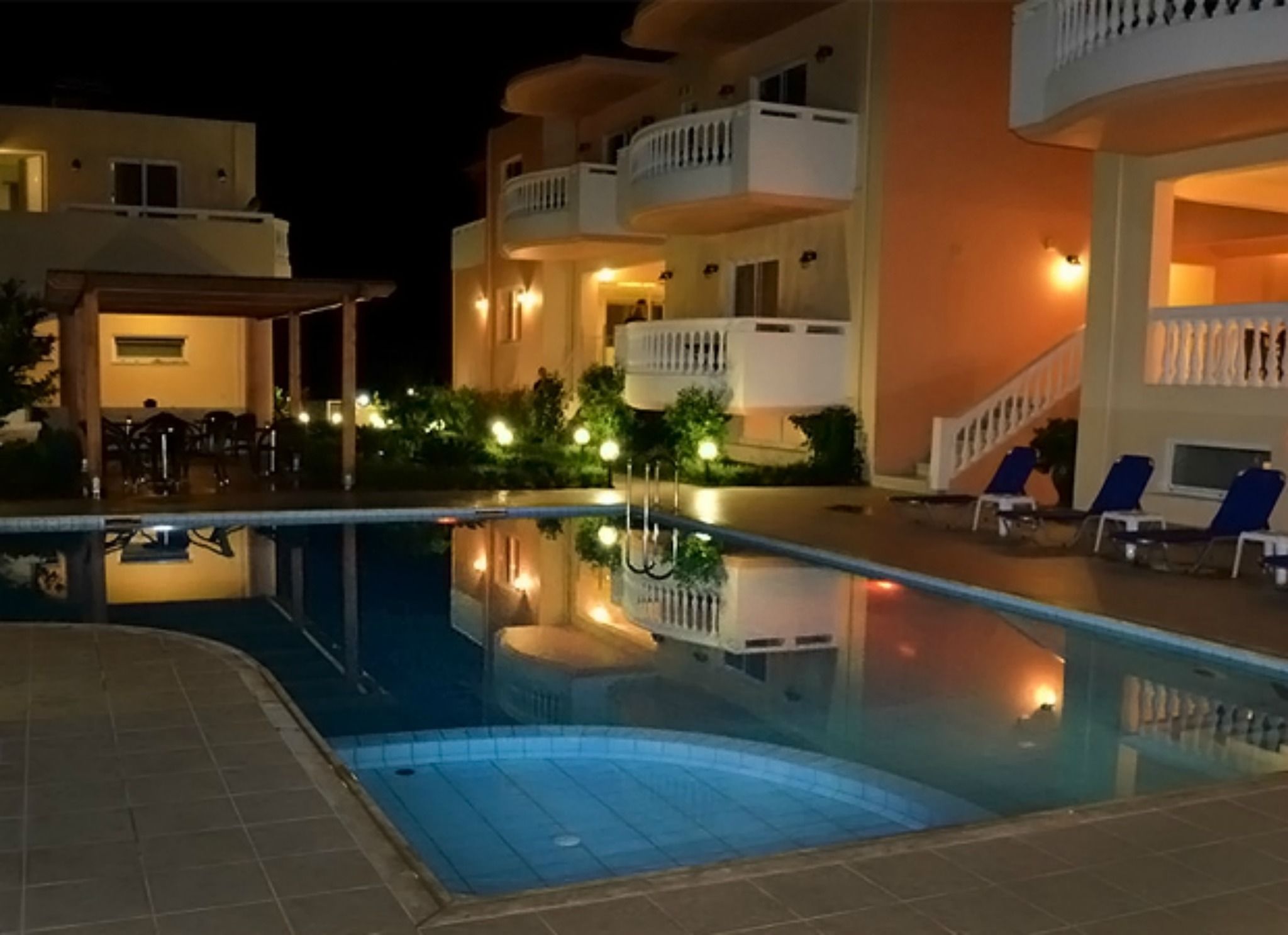 Una piscina con il suo riflesso, circondata da sdraio e un edificio con balconi.