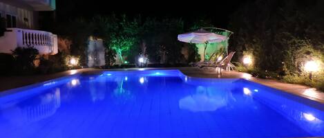 A swimming pool at night with blue lighting, surrounded by trees and a building with balconies.